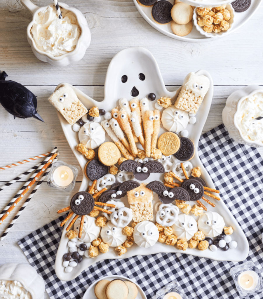 A ghost-shaped platter filled with Halloween treats for kids offers snacks like cookies, pretzels, and popcorn styled as mummies, ghosts, and spiders. It’s accompanied by candles and a black bird decoration on a white wooden table adorned with a checkered cloth.